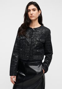 Μαύρο σακάκι με παγιέτες, στρογγυλή λαιμόκοψη, δύο τσέπες στο στήθος και κουμπιά. Συνδυάζεται με μαύρη δερμάτινη φούστα.