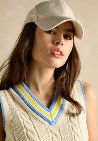 Kreminės spalvos megzta liemenė su kabelių raštais, turinti mėlyną, geltoną ir šviesiai mėlyną banguotą v-kaklą. Šviesiai smėlio spalvos kepurė su herringbone tekstūra.