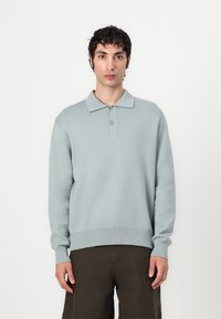 Light blue knitted polo sweater with three-button placket, long sleeves, and ribbed hem. Paired with dark, relaxed-fit trousers.
