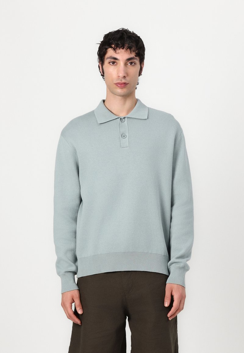 Light blue knitted polo sweater with three-button placket, long sleeves, and ribbed hem. Paired with dark, relaxed-fit trousers.