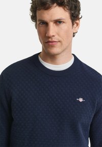 Mörkblå tröja med strukturerat mönster, rundad halsringning och liten logotyp på bröstet. Tillverkad av mjukt tyg med en snug passform.