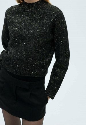 Femme portant un pull en tricot noir moucheté à col haut et une mini-jupe noire, debout les mains dans les poches de la jupe.