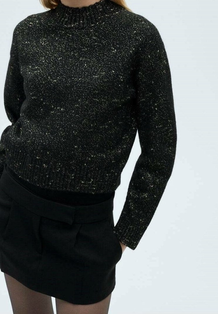 Femme portant un pull en tricot noir moucheté à col haut et une mini-jupe noire, debout les mains dans les poches de la jupe.