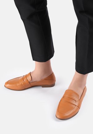 Mocassins en cuir tan avec un embout rond, dotés d'une sangle décorative sur le dessus et d'un petit talon en bois. Portés avec un pantalon noir.