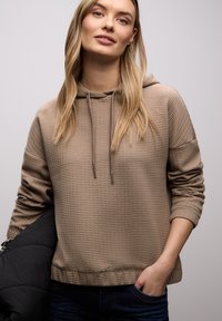 Jeune femme aux longs cheveux blonds portant un sweat à capuche beige texturé et un jean foncé, tenant une veste noire pliée sur un bras.