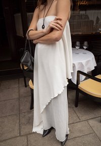 Biała asymetryczna sukienka z falbaną, w połączeniu z czarną torbą i szpilkami za kostkę. Gładki materiał, bezrękawny fason.