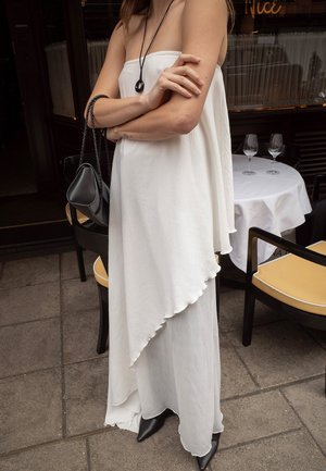 Biela asymetrická šaty s volánkovým lemom, v kombinácii s čiernou kabelkou a špicatými členkovými čižmami. Hladká látka, bezramienkový strih.