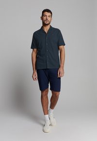 Man in een donkerblauwe, korte mouw, knoopsluiting overhemd, marineblauwe shorts, witte sokken en witte sneakers, staand tegen een effen grijze achtergrond.