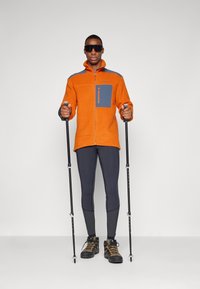 Orange fleece jacket with a gray chest pocket, zip closure, and high collar. The model wears black leggings and hiking boots, holding trekking poles.