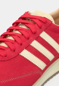 Červená semišová teniska s krémovými pruhmi. Má štruktúrované šnúrky a gumovú podrážku. Ozdobená béžovými detailmi pri jazyku a prešívaní.