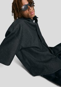 Fekete, oversized nyakkivágású ing, texturált anyagból készült, rövid ujjú és laza szabású. Sötét farmernadrággal és napszemüveggel viselve.