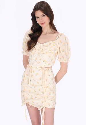 Nuori nainen, jolla on pitkät ruskeat hiukset ja päällä kerma värinen kaksi-osainen asu, jossa on liehukevät hihat ja keltainen kukkaprintti, poseeraa käsillään selän takana.