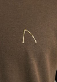 Smeđi kratki rukav majica od mekog pamuka. Karakterizira je minimalistički svijetlosivi dizajn koji na prednjoj strani nalikuje na luk.