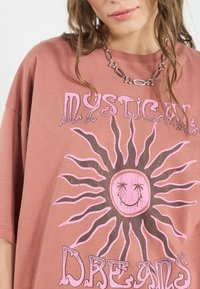 Overdimensioneret rustfarvet T-shirt med et solgrafik i pink og brun. Teksten siger "Mystical Dreams." Glat tekstur, afslappet design.