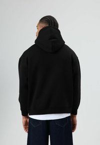 Zwarte hoodie met een grote, ronde capuchon en een losse pasvorm. Heeft geribbelde manchetten en zoom, gedragen over een wit shirt, met een ontspannen silhouet.