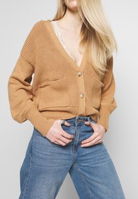 Gestrickte Strickjacke in Kamelbraun mit V-Ausschnitt, zwei aufgesetzten Taschen und Holzknöpfen. Wird über einem Spitzen-Top und hoch taillierten Jeans getragen.