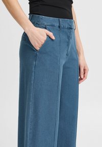 Blauwe wide-leg broek met een gladde textuur, voorzien van een zijzak en een aansluitende taille. De stof heeft een spijkerstofachtige uitstraling.
