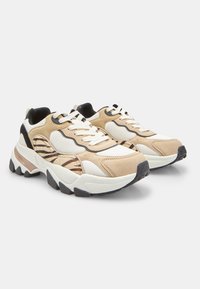 Zapatillas gruesas de color beige y blanco con paneles laterales de estampado animal, cordones blancos, detalles en negro y suelas gruesas de goma negra sobre fondo blanco.