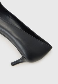 Scarpa con tacco a stiletto nera in pelle vista di lato su uno sfondo chiaro e semplice.