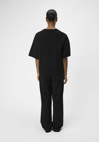 Zwarte oversized top met korte mouwen en een textuurafwerking, gecombineerd met losse zwarte broek, gezien vanaf de achterkant tegen een lichte achtergrond.