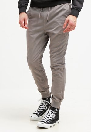 Wąskie, jasnoszare spodnie dresowe ze ściągaczami, noszone z czarnymi trampkami za kostkę z białymi sznurówkami.