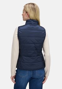 Marineblaues ärmelloses Weste, gestepptes Design, hoher Kragen, leichtes Material. Trägt man über einem cremefarbenen Pullover, kombiniert mit blauen Jeans.