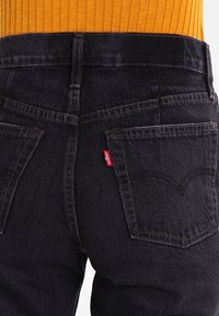 Jeans de mezclilla negra de cintura alta, con bolsillos traseros y un logo en forma de etiqueta roja. La tela tiene una textura suave y costuras prominentes.