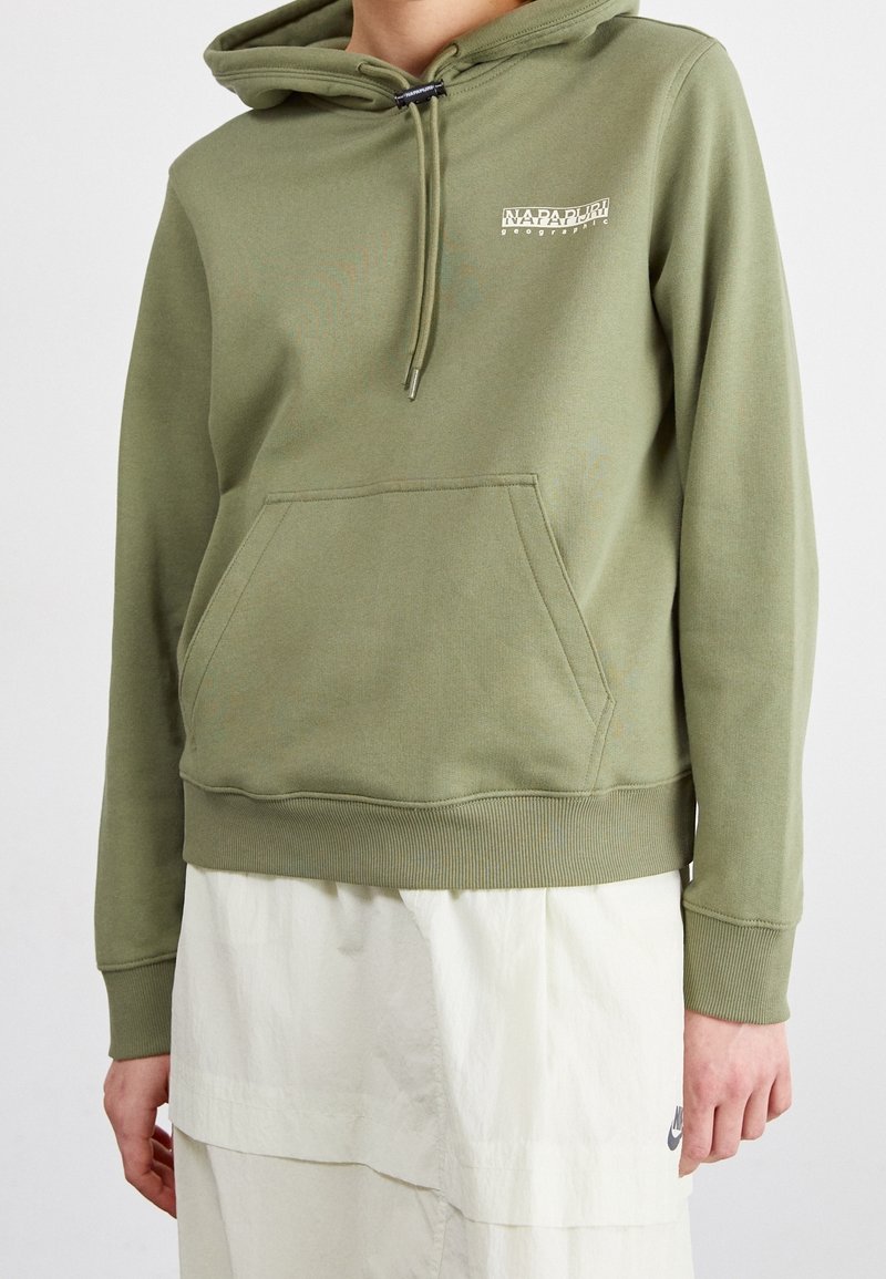 Personne portant un sweat à capuche vert olive avec poche avant et logo texte blanc, associé à une jupe blanche à cordon avec un petit logo Nike.