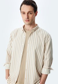 Camisa de botones a rayas beige con una textura suave, con cuello y mangas remangadas, sobre un suéter ligero beige.