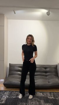 Γυναίκα που φοράει μαύρο t-shirt και παντελόνι στέκεται ξυπόλυτη σε ένα σαλόνι με γκρι δερμάτινο καναπέ και χαλί με σχέδια πίσω της.