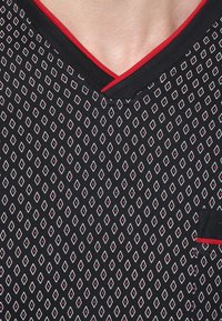 Schwarzer Stoff mit einem Diamantmuster in Weiß und Rot. V-Ausschnitt-Design mit roten Akzenten entlang des Ausschnitts und einer Tasche mit rotem Rand.