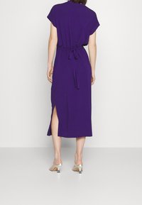 Fialové midi šaty s krátkými rukávy, detailem na zavázání v pase a bočním rozparkem. Hladká látka s mírným splývavým efektem, doplněná o transparentní sandály na podpatku.