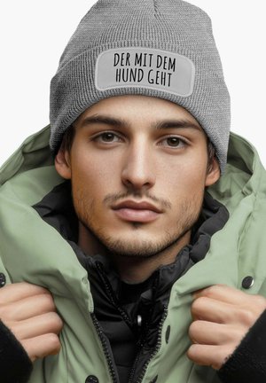 Gray knit beanie with text patch "DER MIT DEM HUND GEHT" worn with a green puffer jacket over a black layer, showcasing a layered look.