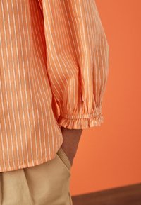 Light orange striped shirt with white lines, featuring puffed sleeves and elastic cuffs. Worn with beige trousers on a contrasting background.