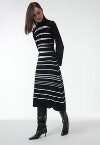 Zwart-wit gestreepte gebreide jurk met hoge hals en lange mouwen; het aansluitende bovenlijf gaat over in een uitlopende rok; gecombineerd met zwarte puntige laarzen.