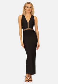 Schwarzes Abendkleid mit einer taillierten Silhouette, tiefem V-Ausschnitt, transparentem Einsatz an der Taille und langem, schmalem Design. Getragen mit schwarzen Riemchenschuhen.