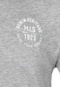 Šedé bavlněné tričko s kulatým bílým logem, které nese nápis "DENIM HERITAGE H.I.S 1923 NEW YORK BRAND" v tučném písmu.