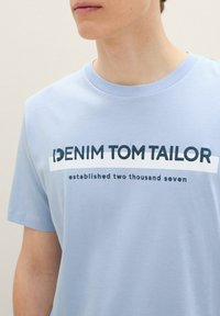 Šviesiai mėlynas medvilninis marškinėlis su apvaliu iškirpimu ir trumpomis rankovėmis. Priekinis grafinis vaizdas apima užrašą "DENIM TOM TAILOR" ir "įsteigtas 2007 metais" tamsiai mėlyna spalva.