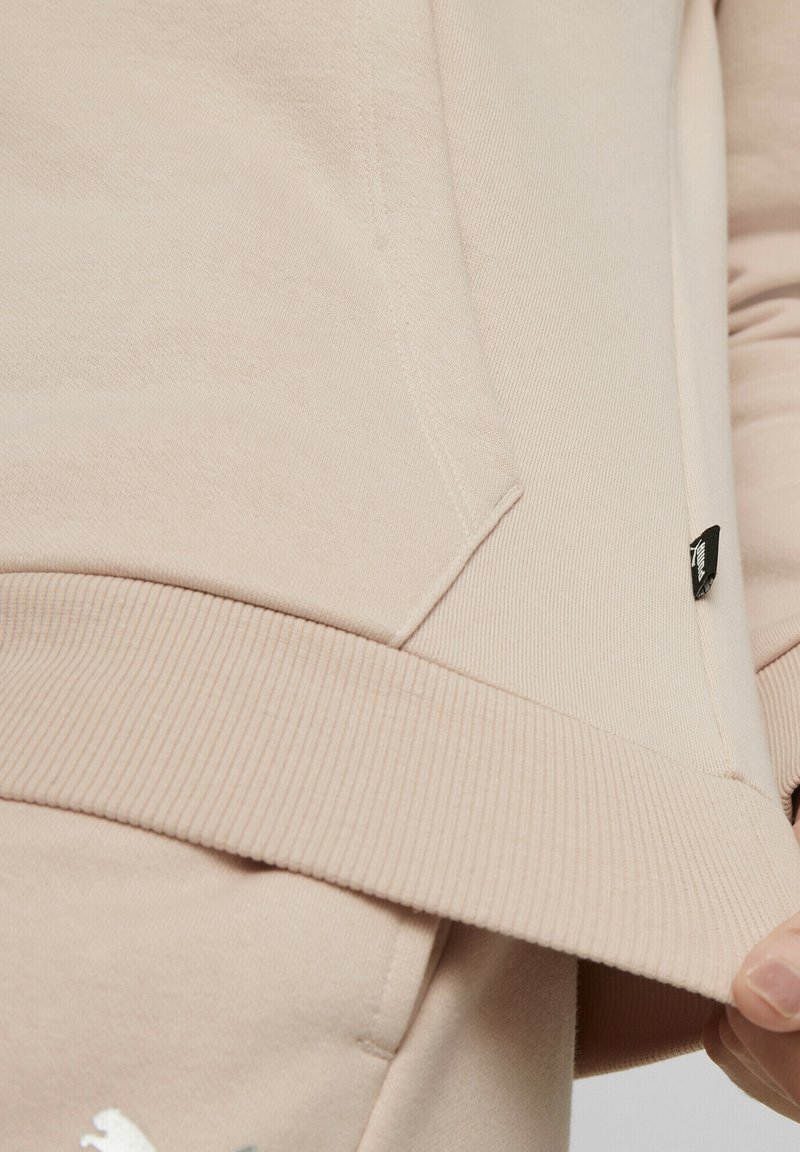 Gros plan sur un sweat-shirt beige montrant un ourlet côtelé, une poche avant, une petite étiquette noire avec le logo, et une partie de la main soulevant l'ourlet du vêtement.