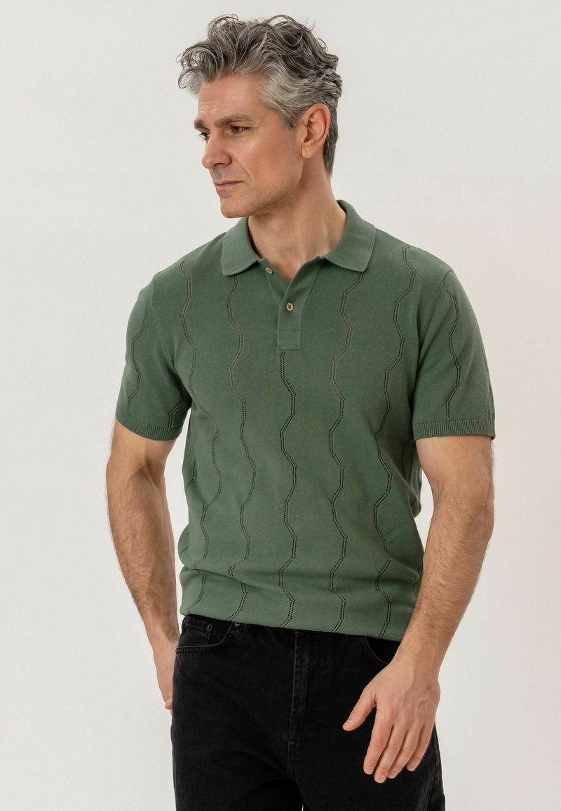 Homme aux cheveux poivre et sel portant un polo à manches courtes vert à motifs et un pantalon noir, regardant sur le côté les mains détendues.