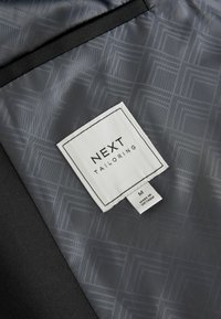 Label til et sort beklædningsgenstand med grå, geometrisk mønsterforing. Tag viser "NEXT TAILORING" og størrelse "M," lavet i Vietnam.