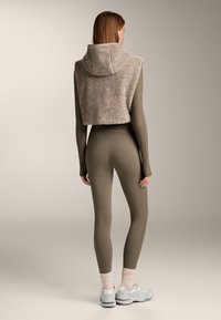 Rövidített bézs bolyhos kapucnis pulóver, zöld ribbelős bodyn és azonos árnyalatú leggings alatt viselve, világosszürke sportcipőkkel párosítva.