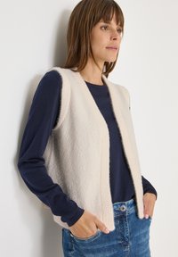 Beige gestrickte Weste mit offenem Design, getragen über einem navyfarbenen Langarmshirt. Hat eine weiche Textur und eine lässige Passform.