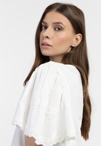 Hvid, let bluse med puffede ærmer, delikat blonder trim og en tekstureret overflade. Modellen vises fra siden med hår, der falder ned.