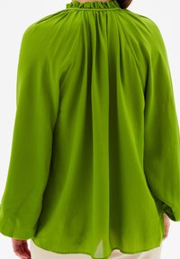 Blusa verde con un colletto alto e arricciato, maniche lunghe e tessuto morbido e fluido. Presenta pieghe sul retro. Texture leggera e ariosa.