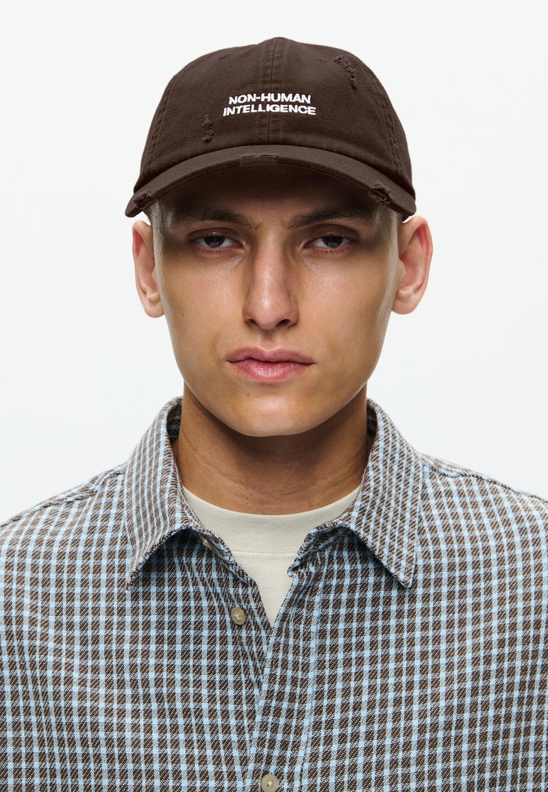 Cappello di cotone marrone con la scritta "NON-HUMAN INTELLIGENCE" ricamata in bianco. Il portatore indossa una camicia a quadri nei toni di marrone e blu.