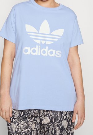 Personne portant un T-shirt Adidas bleu clair avec un logo et un texte blanc, associé à un pantalon imprimé serpent noir et beige.