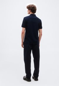 Νεαρός άνδρας με σγουρά μαλλιά στέκεται με την πλάτη, φορώντας μια υφασμάτινη μαύρη κοντομάνικη πόλο, μαύρο παντελόνι και μαύρα παπούτσια σε απλό φόντο.