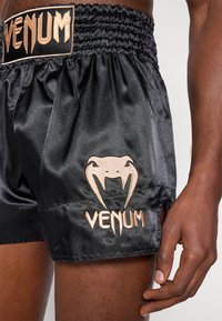 Juodi satino šortai su surinktą juosmenį, puoštas auksinio siuvinėjimo „VENUM“ tekstu ir logotipu bei šoninėmis iškirpomis, suteikiančiomis daugiau judesio.