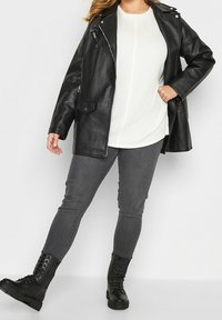 Černá faux kožená bunda se zipovými a klopovými kapsami přes bílý žebrovaný top, spárovaná se šedými skinny džíny a černými šněrovacími botami.